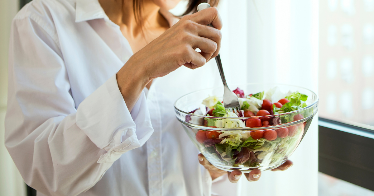 Close-up of a woman in a white shirt holding a fork and a glass bowl filled with fresh salad, including cherry tomatoes and mixed greens.