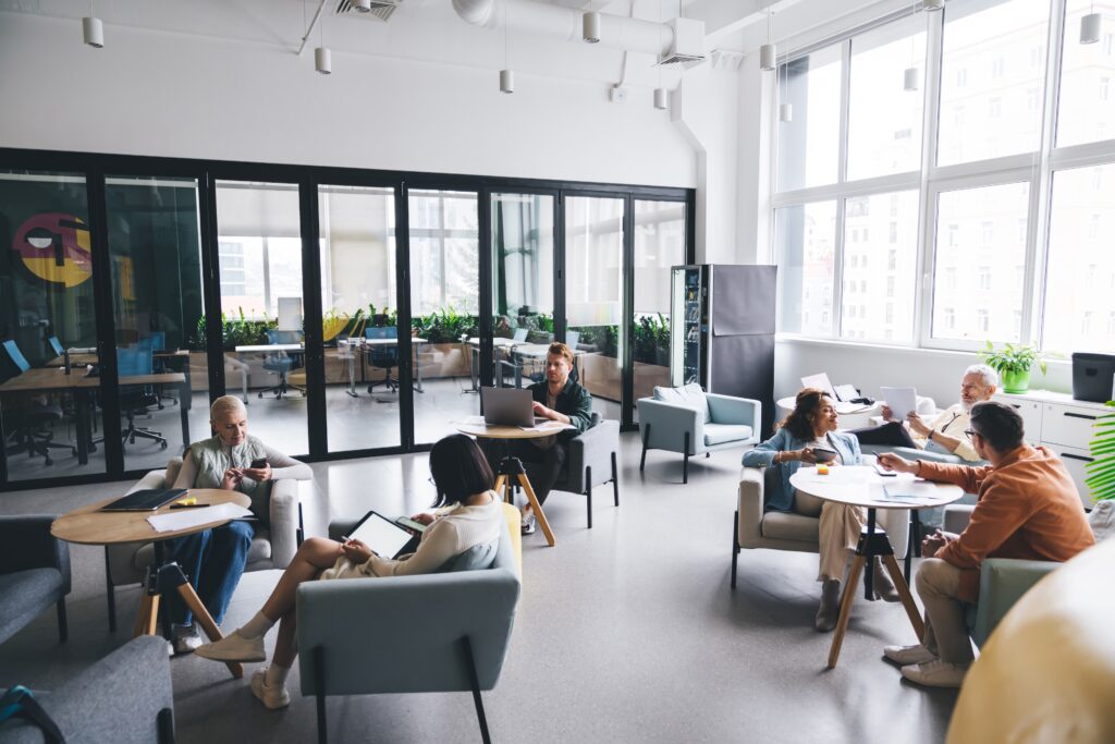 AdobeStock 577440202 2 A modern, open-concept coworking space with people engaged in various activities, including working on laptops, reading documents, and having discussions. The space is well-lit with large windows, comfortable seating, and greenery.