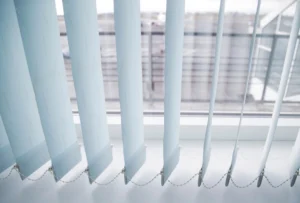 Blinds 1 This image shows a close-up view of vertical window blinds, partially open, with a view of the outside. The blinds are light blue and have a chain linking them at the bottom. The window provides a glimpse of an exterior scene with blurred buildings and structures, allowing natural light to filter through and illuminate the blinds. The overall feel is clean and modern.