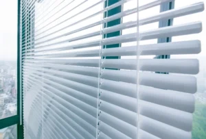 Blinds 2 This image depicts a close-up view of horizontal window blinds, partially open to allow a clear view of the outside. The blinds are white and positioned in front of large windows that offer a glimpse of a cityscape with buildings and greenery. The natural light filtering through creates a bright and airy atmosphere, emphasising the clean and modern design of the space.
