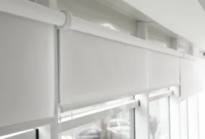 Blinds 3 This image shows a close-up view of roller blinds in a modern setting. The blinds are white and fully extended, covering the windows. A pull chain for adjusting the blinds hangs on the side. The windows provide a faint, blurred view of the outside, allowing natural light to softly filter into the room. The overall design is clean and minimalist, enhancing the contemporary feel of the space.