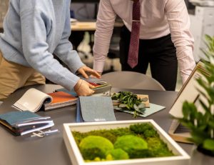 Two individuals are engaged in a design discussion, examining fabric swatches and colour samples spread out on a table. One person is wearing a light blue sweater, while the other is in a pink shirt with a red tie. The table also features a small tray with greenery, adding a natural touch to the workspace. The background is slightly blurred, highlighting the focus on the materials and the collaborative activity.
