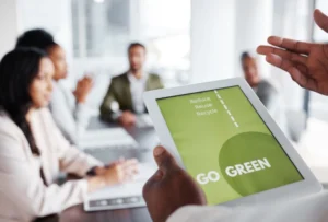 A team of professionals is in a meeting, with the focus on a person holding a tablet displaying a "Go Green" message. The tablet screen reads "Reduce, Reuse, Recycle" in green text, emphasising sustainability. The background shows blurred colleagues in discussion, highlighting the modern, collaborative office environment. The setting is bright and professional, with natural light coming through large windows. 