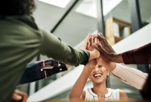 A group of diverse colleagues enthusiastically perform a group high-five in a modern office setting. The focus is on their hands meeting in the centre, symbolising teamwork and unity. A smiling woman is visible in the background, enhancing the positive and collaborative atmosphere. The office features glass walls and contemporary design elements.